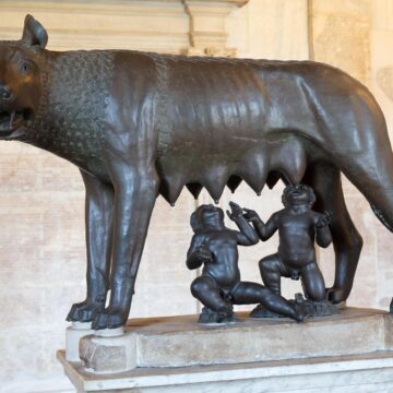 Inside Roman Mythology: Greek Gods, Roman Ambitions Bronze sculpture of a she-wolf nursing two infants on a stone pedestal.