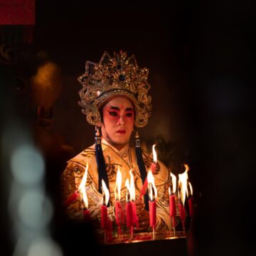 Chinese Mythology: A Giant’s Body Becomes the World Chinese opera performer in an ornate gold headdress and embroidered costume stands behind burning candles.