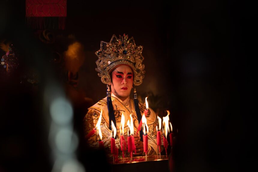 Chinese opera performer in an ornate gold headdress and embroidered costume stands behind burning candles.