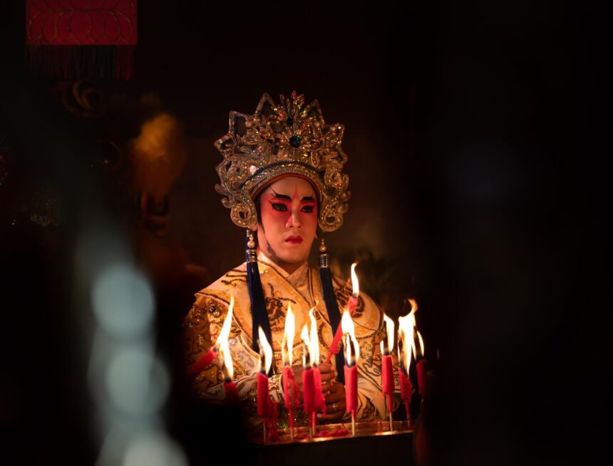 Chinese opera performer in an ornate gold headdress and embroidered costume stands behind burning candles.