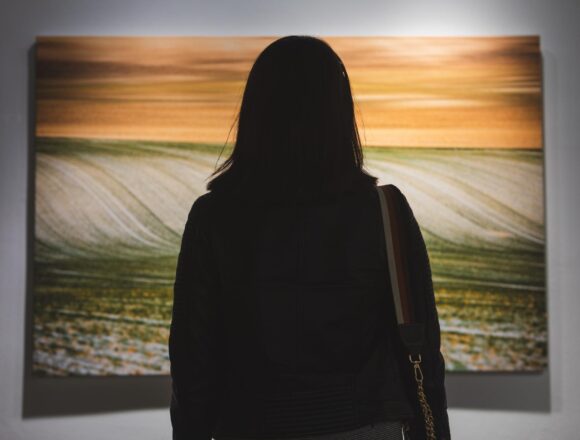 A woman views a large painting of rolling fields beneath an orange sky in a gallery.