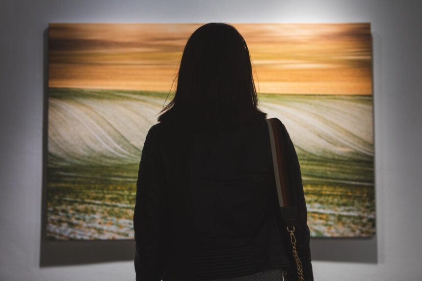 A woman views a large painting of rolling fields beneath an orange sky in a gallery.