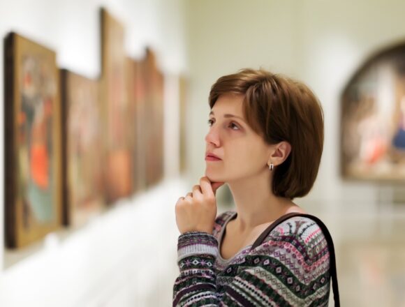 A visitor studies a row of framed paintings in a softly lit art gallery.