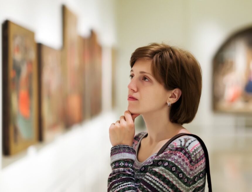 A visitor studies a row of framed paintings in a softly lit art gallery.