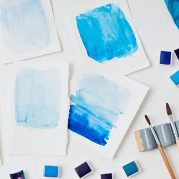 14 Famous Watercolor Paintings: Sharp Edges, Soft Washes Blue watercolor washes on sheets of paper beside paint pans and brushes on a white surface.