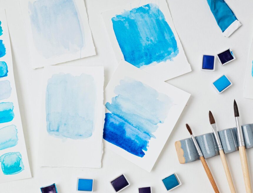 14 Famous Watercolor Paintings: Sharp Edges, Soft Washes Blue watercolor washes on sheets of paper beside paint pans and brushes on a white surface.