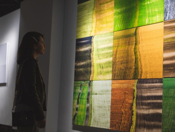 A woman studies a large abstract painting with green, yellow, orange and dark rectangular blocks in a gallery.
