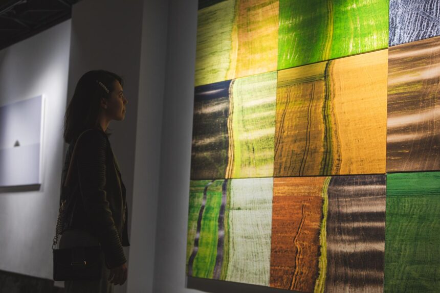 A woman studies a large abstract painting with green, yellow, orange and dark rectangular blocks in a gallery.