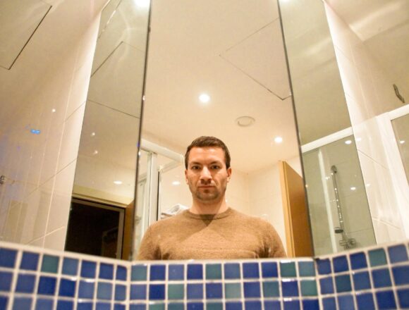 A man's reflection in a bathroom mirror above a row of blue tiles.