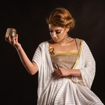 Pandora Greek Mythology: Hesiod’s Jar and Uncertain Hope Woman in a white and gold draped dress holding up a small gold-colored cup against a black background.