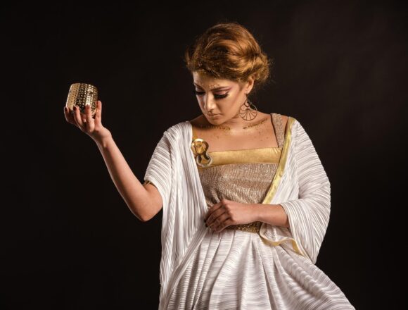 Woman in a white and gold draped dress holding up a small gold-colored cup against a black background.