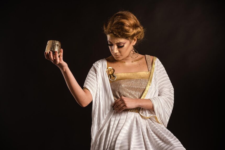 Woman in a white and gold draped dress holding up a small gold-colored cup against a black background.