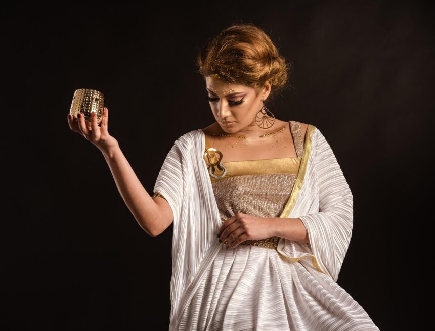 Pandora Greek Mythology: Hesiod’s Jar and Uncertain Hope Woman in a white and gold draped dress holding up a small gold-colored cup against a black background.