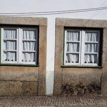 Two curtained windows with stone surrounds sit low above a cobbled pavement on a weathered facade.