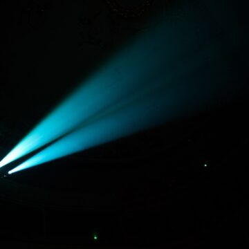 Project Blue Beam: A 1994 Story of Manufactured Miracles Two blue searchlight beams cut through haze in a dark concert hall.