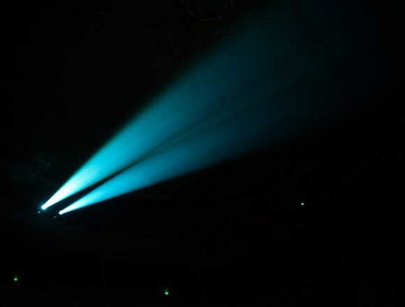 Project Blue Beam: A 1994 Story of Manufactured Miracles Two blue searchlight beams cut through haze in a dark concert hall.