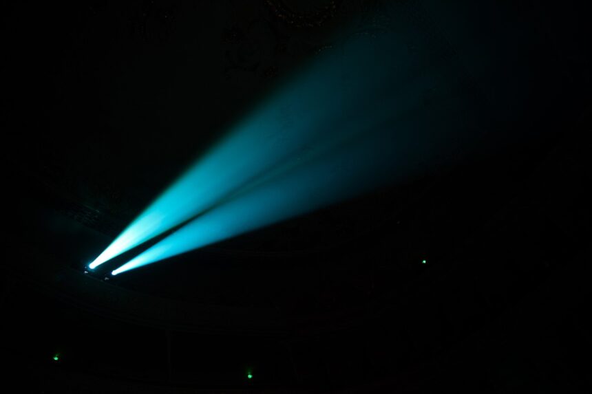 Project Blue Beam: A 1994 Story of Manufactured Miracles Two blue searchlight beams cut through haze in a dark concert hall.
